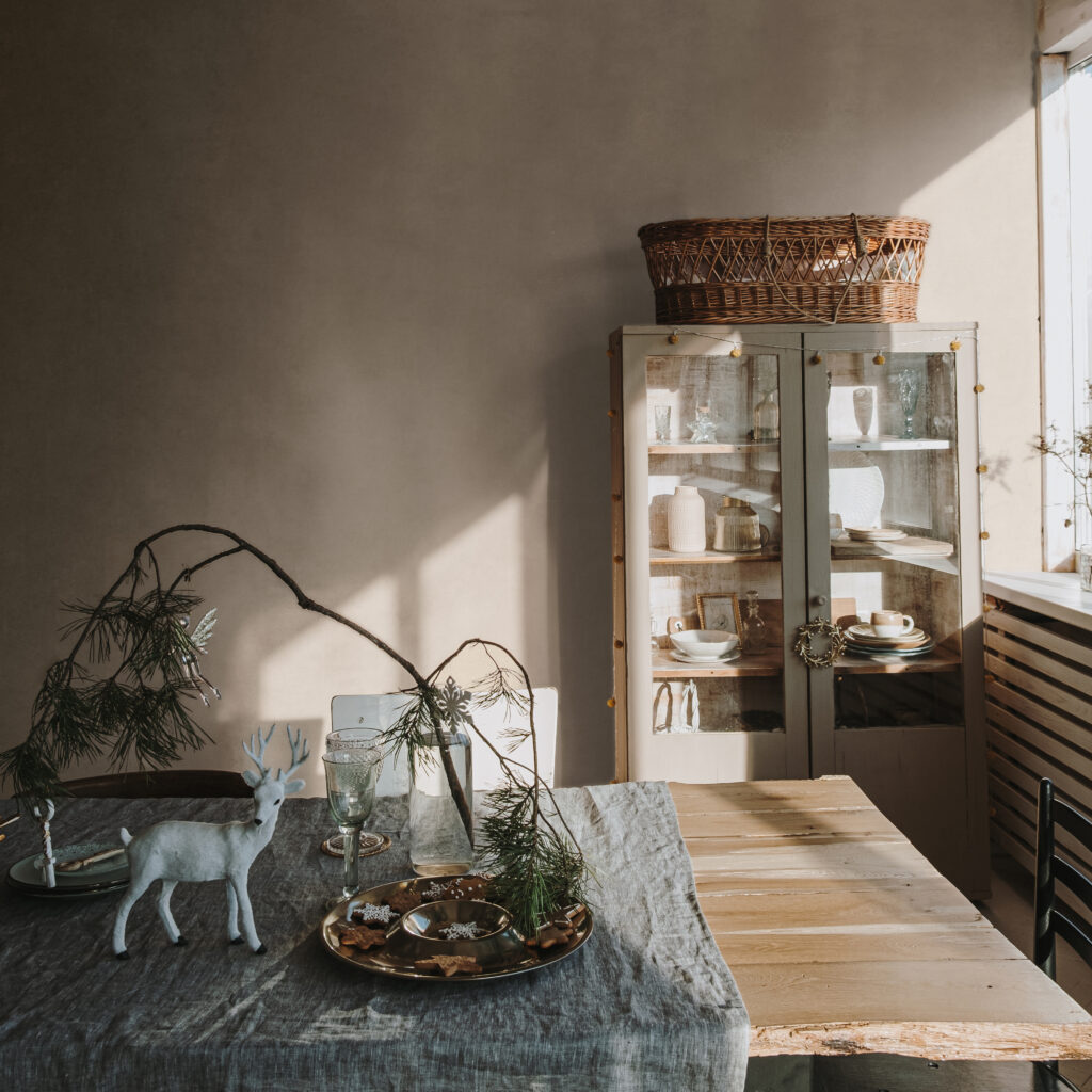 Elegant aesthetic home living or dinning room interior design with dinner table, linen table cloth, fir branch bouquet, Christmas wreath, neutral dusty pink walls. Sunlight shadows on the wall